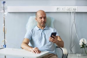 Portrait of adult man using smartphone during IV drip treatment in hospital, copy space
